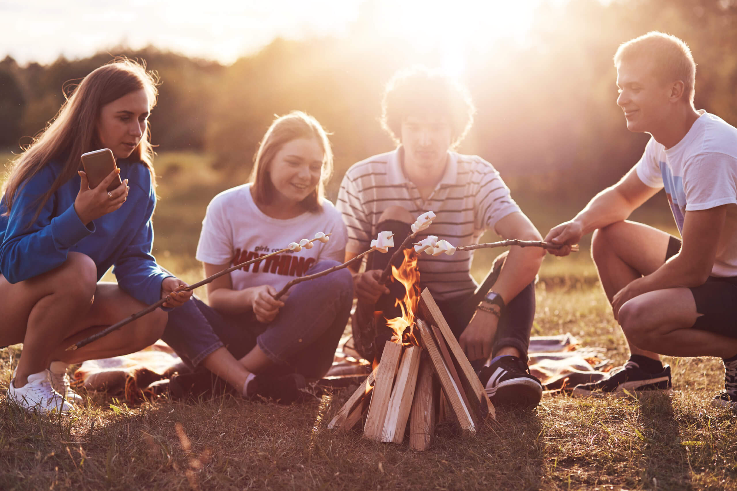 Photo of happy classmates or friends roast marshmallows over campfire, sit on ground, have picnic together during summer day, nice sunny weather, have pleasant talk. Togetherness and lifestyle concept